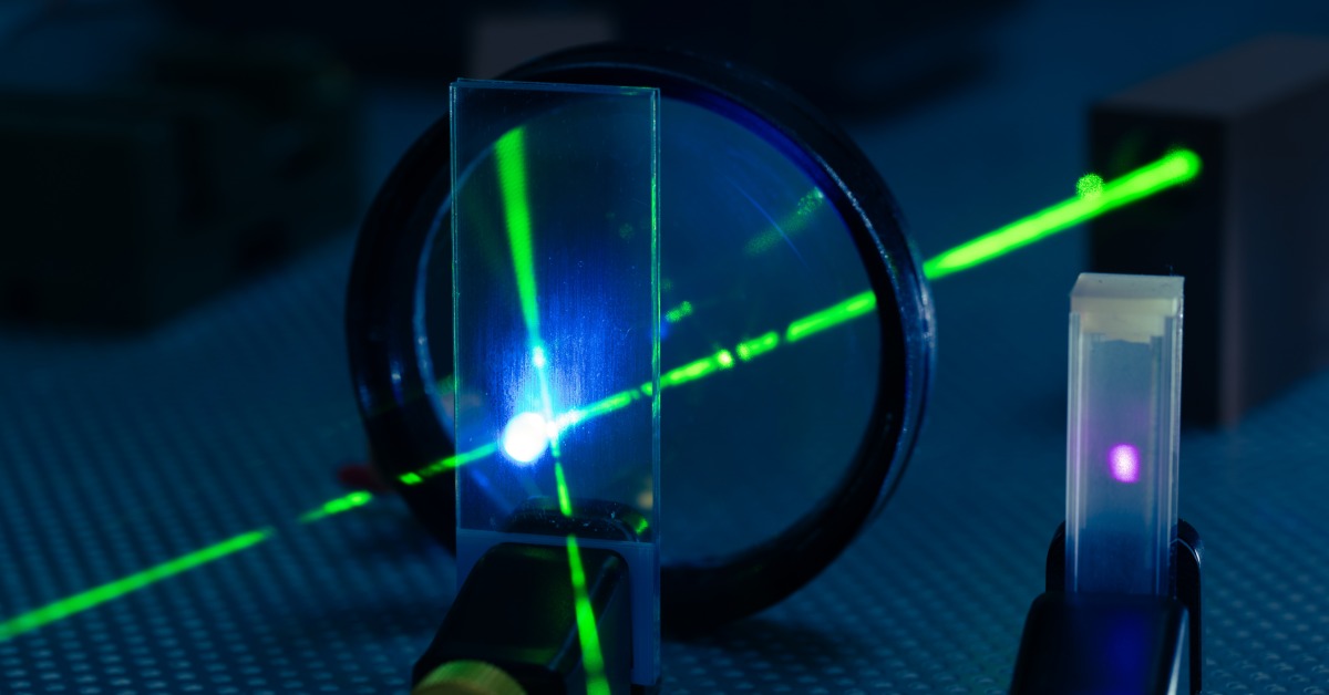 A green laser crosses clear optical components as rectangular glass elements redirect the beam across a dark lab setup.