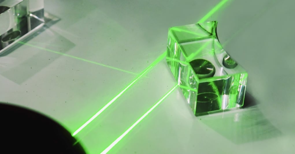 A green laser shines a beam of light into a custom glass component, as it redirects the beam across a white surface.