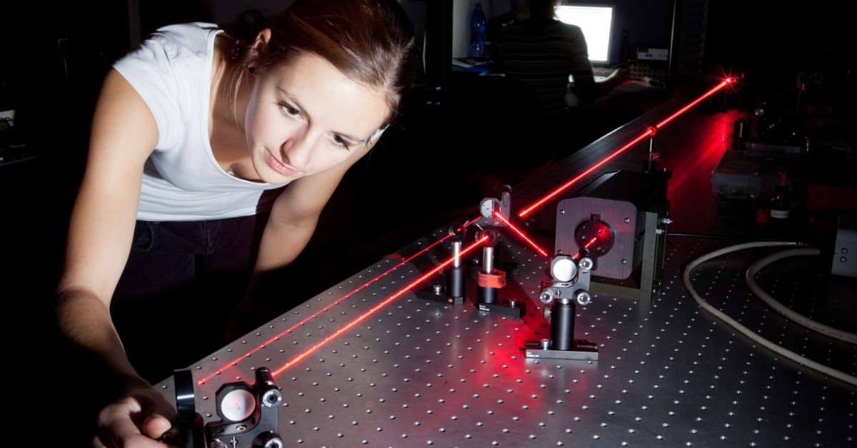 A researcher adjusts a mounted optic while red laser beams travel across an optical table in a dark laboratory.