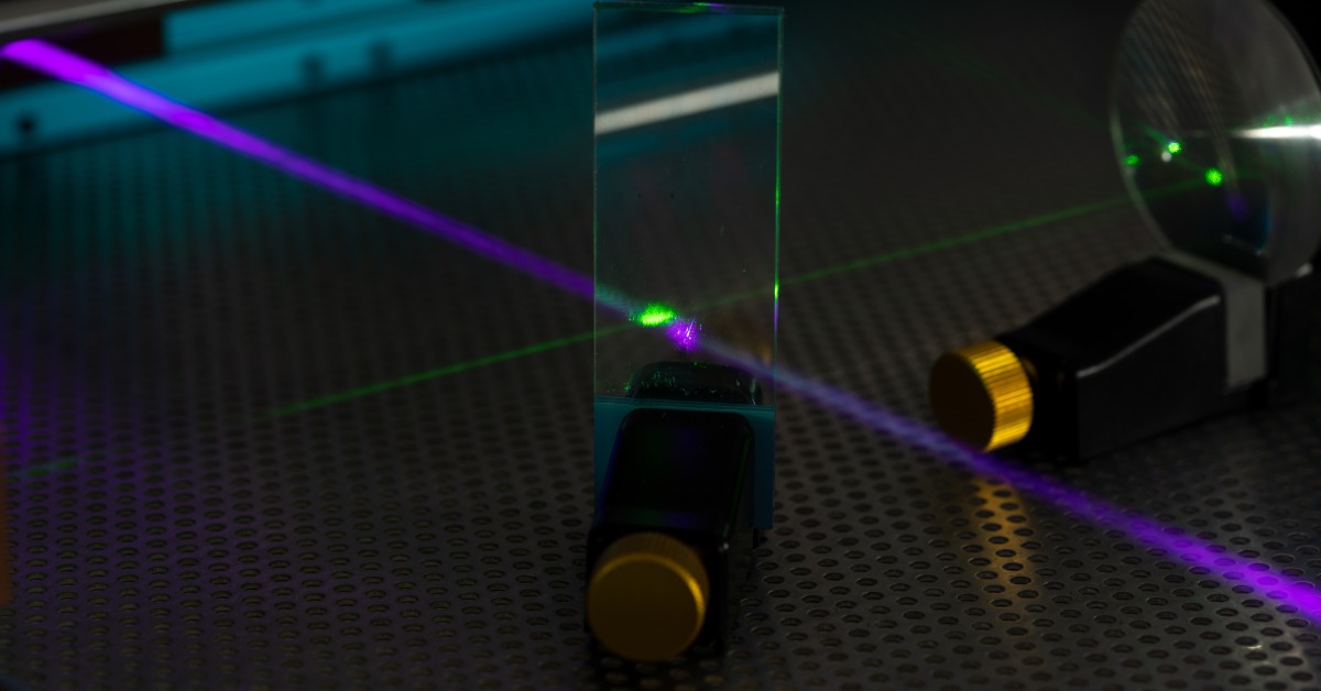 Green and purple laser beams pass through upright glass prisms and optical components in a dark laboratory.