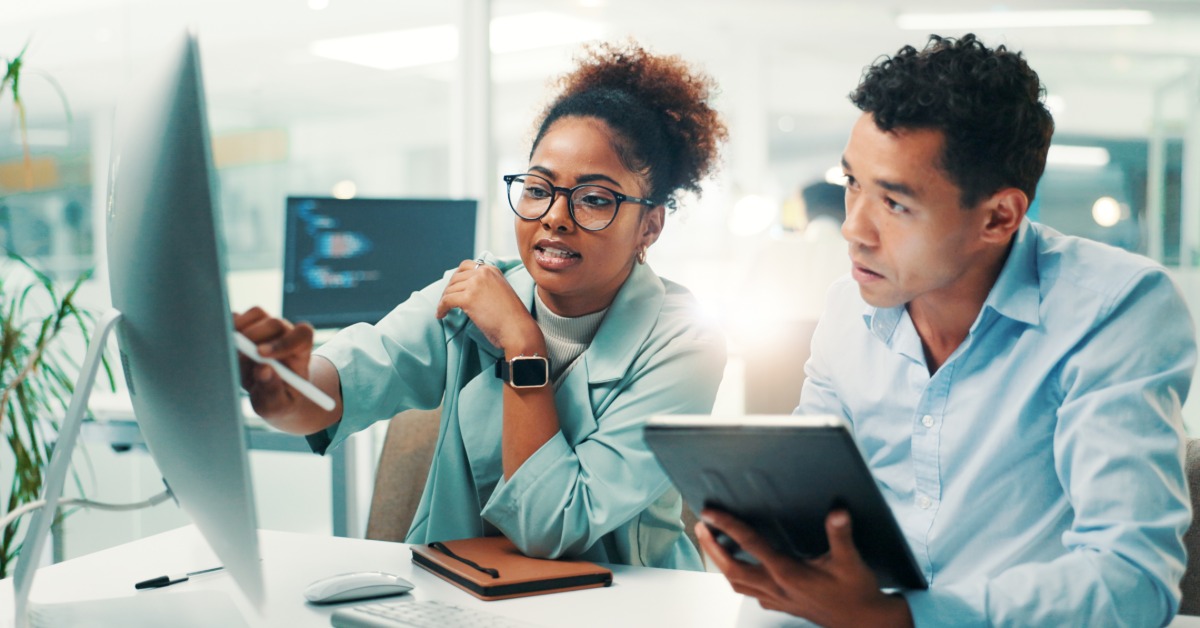 Two professionals review information on a desktop monitor while one points at the screen and the other holds a tablet.