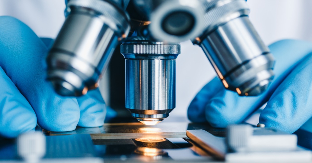 A technician in blue gloves examines a glass slide beneath the metal objective lenses of a laboratory microscope.