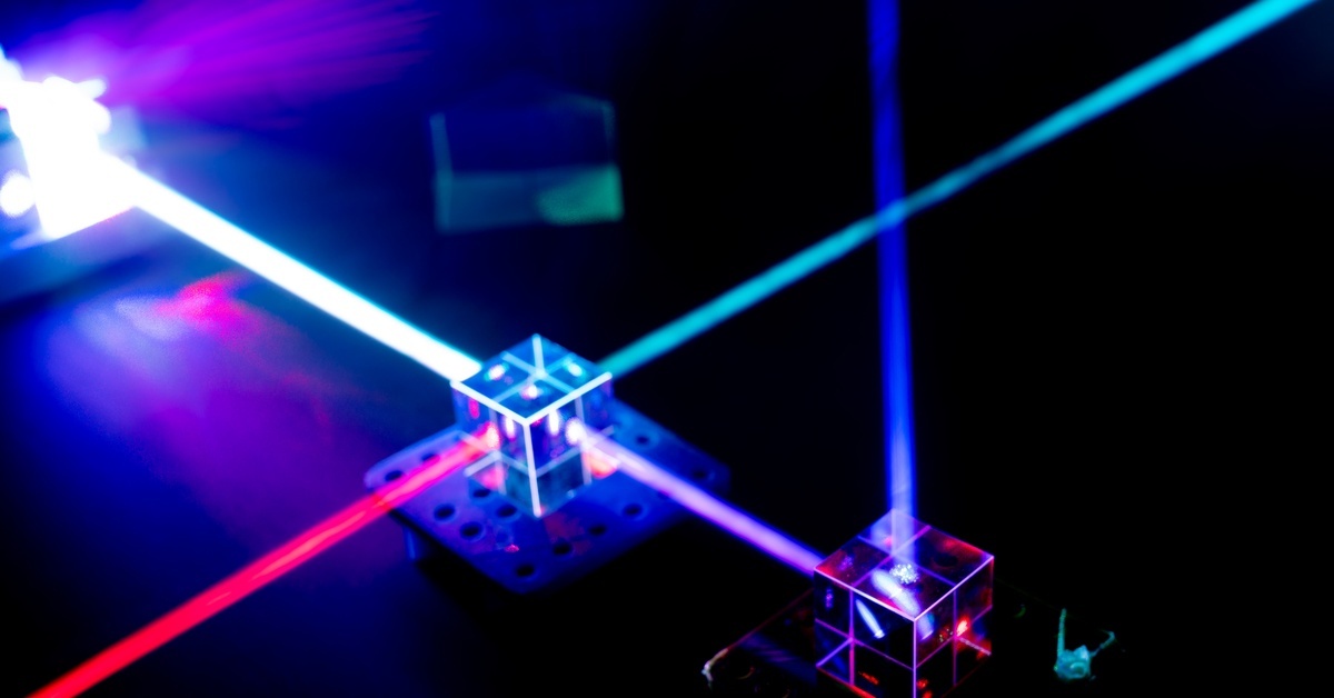 A bright blue laser beam passes through clear cube optics while red and violet beams cross a dark optical table.