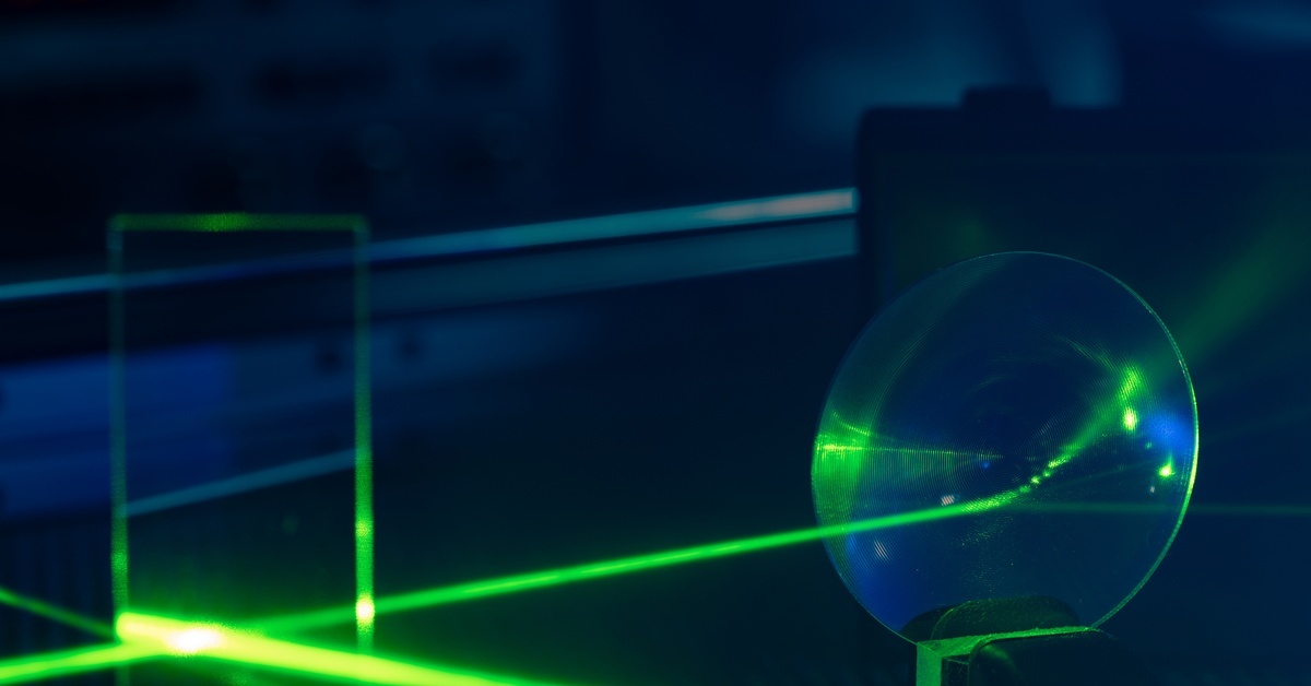A green laser beam strikes a round optical element as other beams cross a dark laboratory optical table.