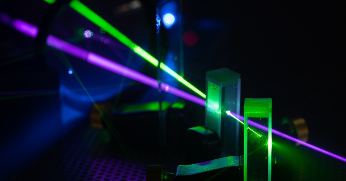 Green and violet laser beams pass through upright glass optics mounted across a dark laboratory optical table.