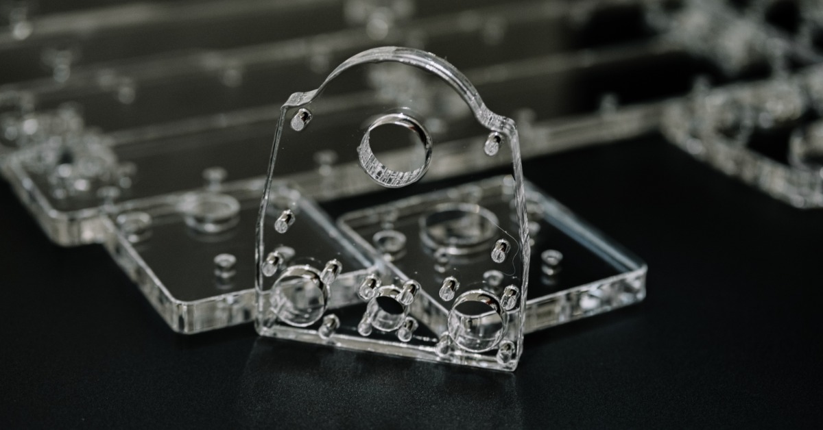 A custom glass component with drilled holes stands in front of matching clear parts on a dark inspection surface.