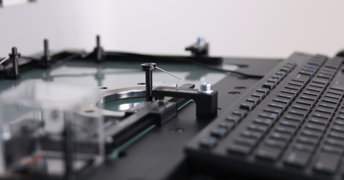A precision inspection machine holds a glass part beside a keyboard and metal fixtures in a technical lab.