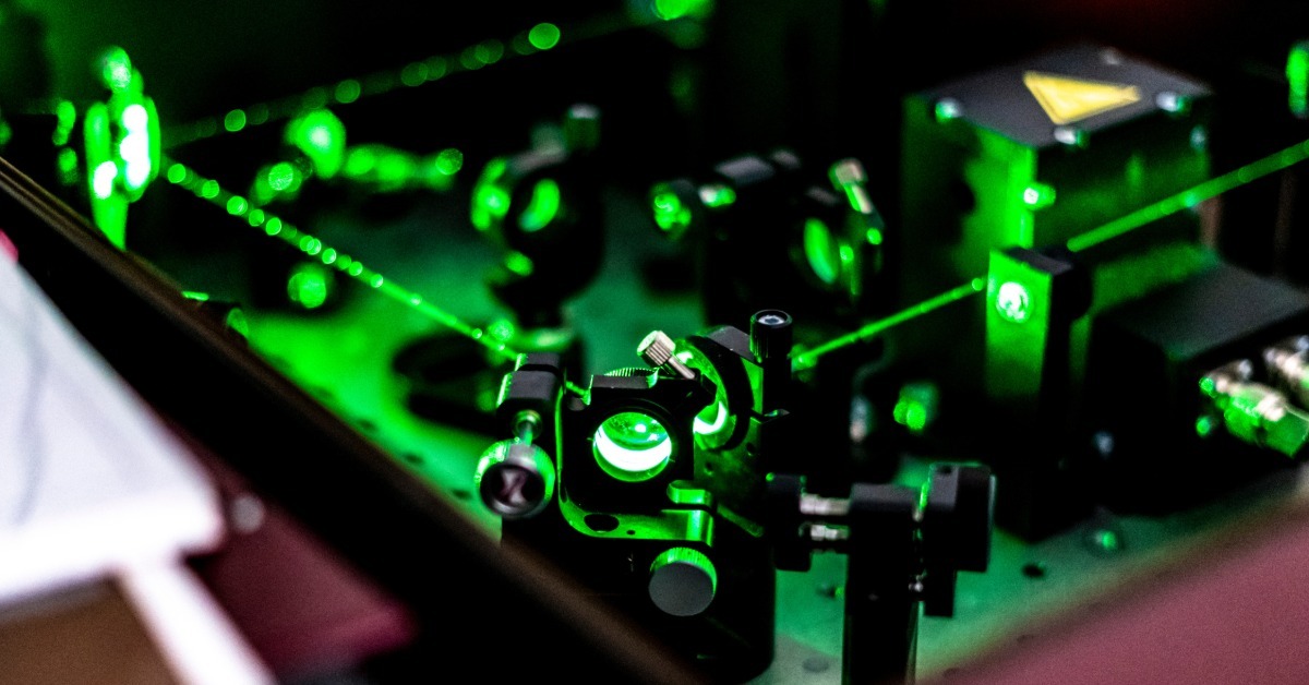 A green laser beam crosses mirrors and mounts on an optical table inside a dark photonics laboratory.