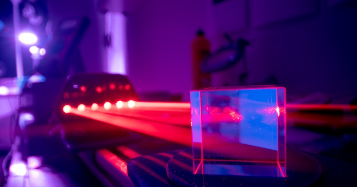 A red laser beam passes through a clear glass block on an optics bench in a dark purple optics laboratory.
