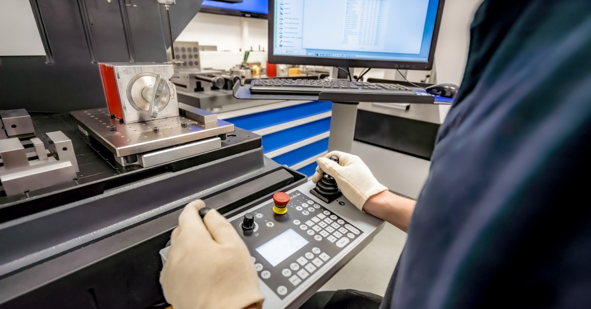 A technician operates a control panel beside a precision machine with a monitor in a manufacturing lab.