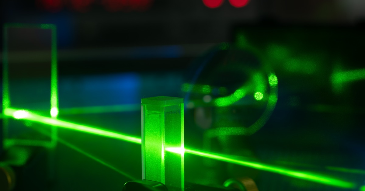 A green laser beam passes through a tall glass block on an optical table as laboratory equipment fills the background.