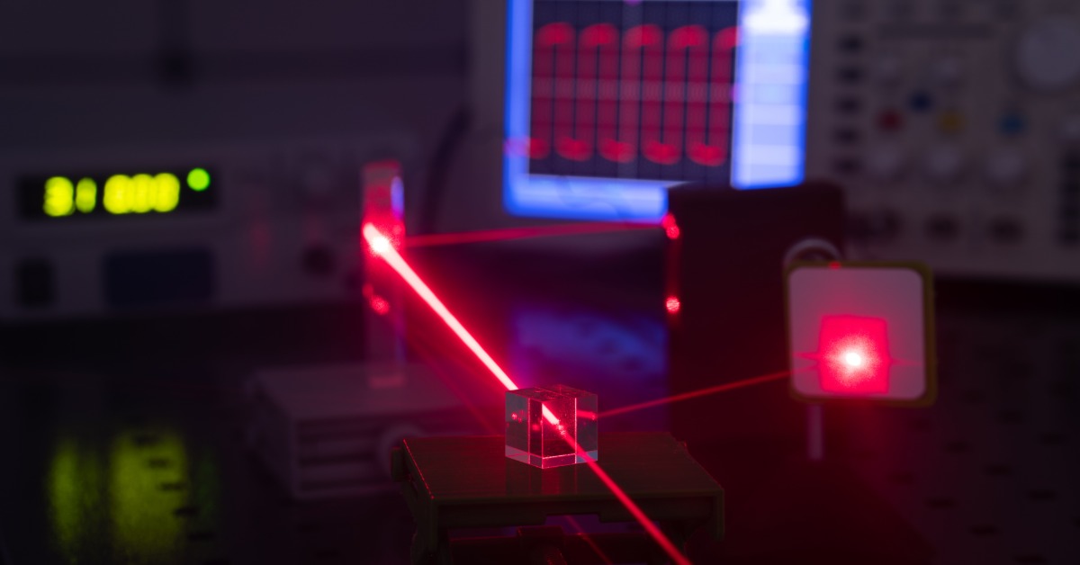 A red laser beam passes through a clear glass cube on an optical table while laboratory instruments glow in the background.